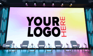 Stage with a large screen reading 'YOUR LOGO HERE' and six gray chairs lined up on stage beneath lighting.